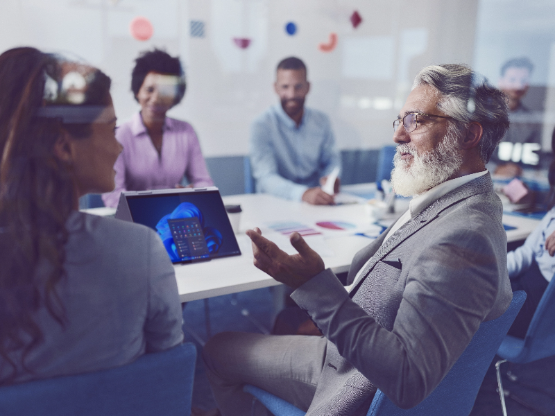 Mature bearded man with laptop with Windows 11 displayed, converses with large group of people at office conference table.