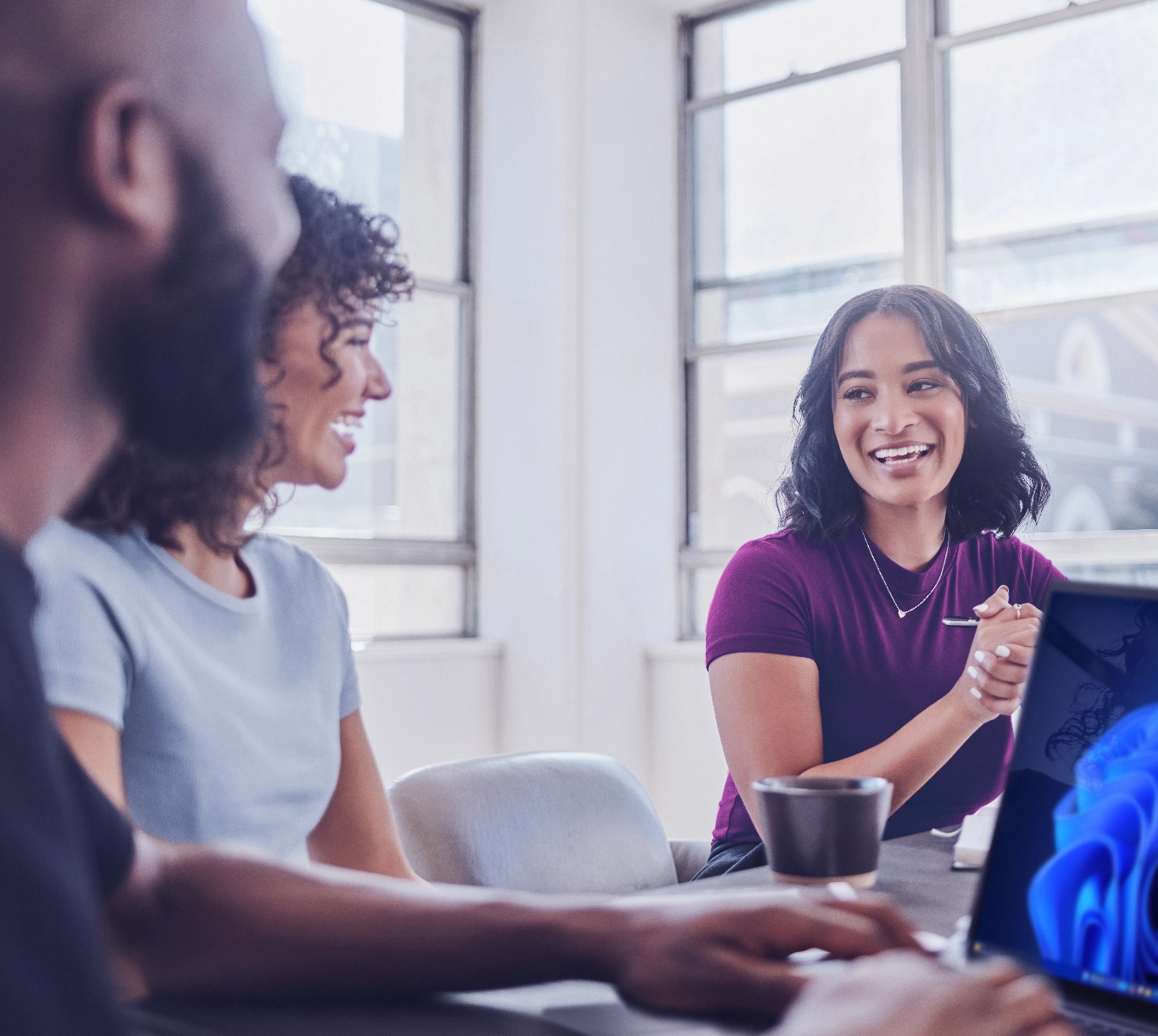 Group of  three people smiling sitting around an office conference table with laptop displaying Windows 11 desktop background.